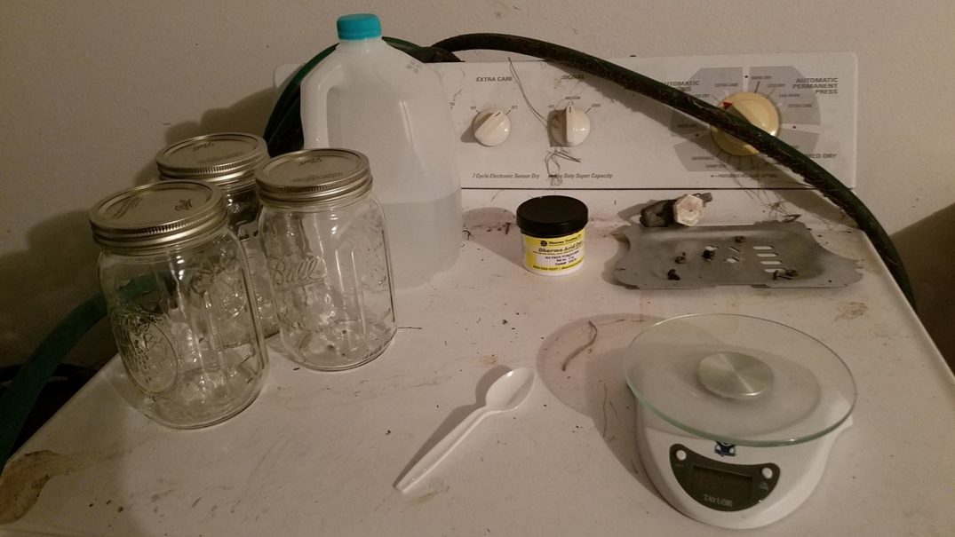 Three pint sized mason jars, a food scale, a plastic spoon, half a gallon of distilled water, and a tub of acid dye sitting on top of a dirty dryer in a garage.